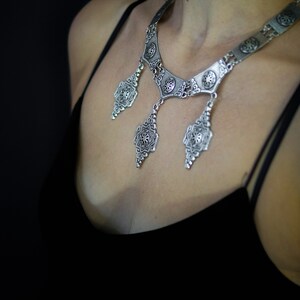 May include: A silver necklace with a geometric design and multiple pendant drops. The necklace is worn by a person with a black top.