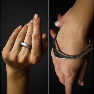 May include: Two silver rings, one a simple band and the other a wide, curved band with an inscription. Both rings are worn on a person's hand.