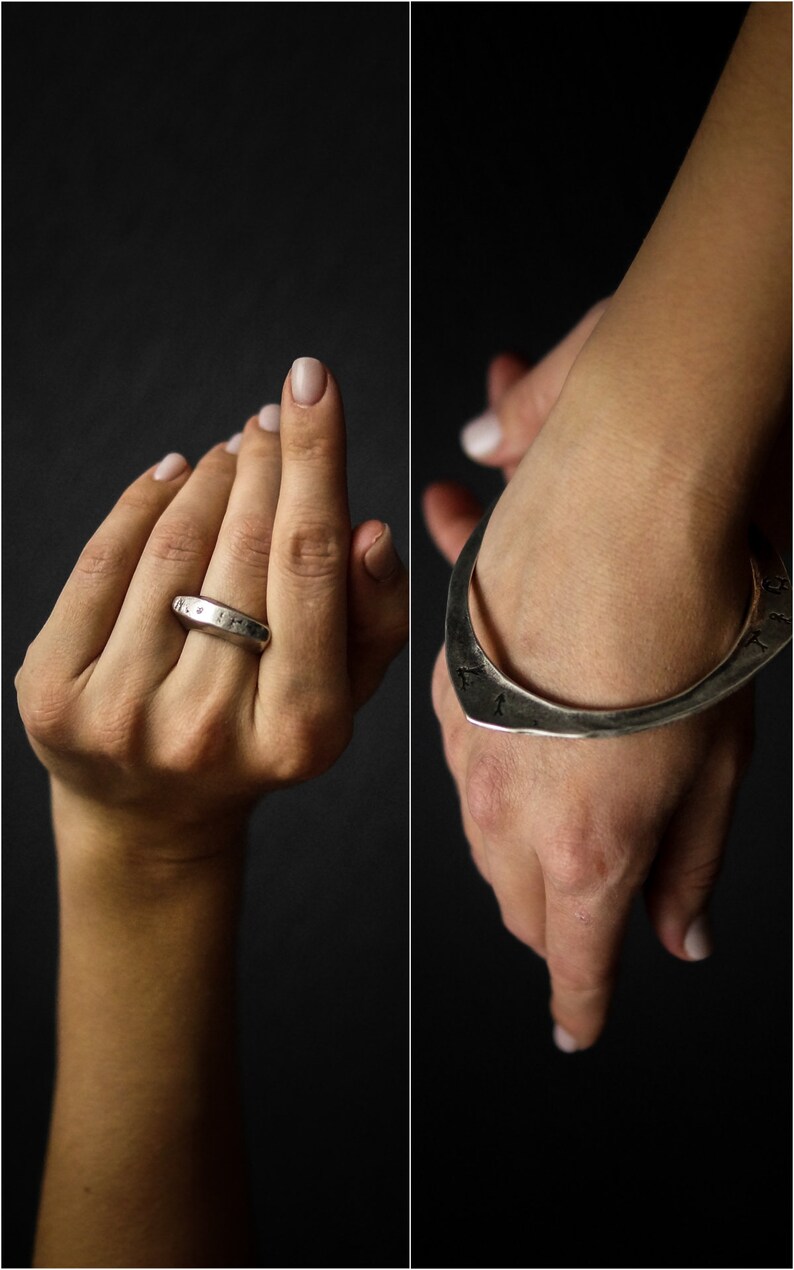 May include: Two silver rings, one a simple band and the other a wide, curved band with an inscription. Both rings are worn on a person's hand.