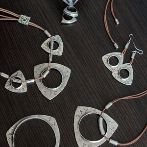 May include: A set of silver jewelry with a triangular pendant design. The pendants feature a circle with intricate engravings and are attached to brown leather cords. The set includes a necklace, earrings, and a bracelet.