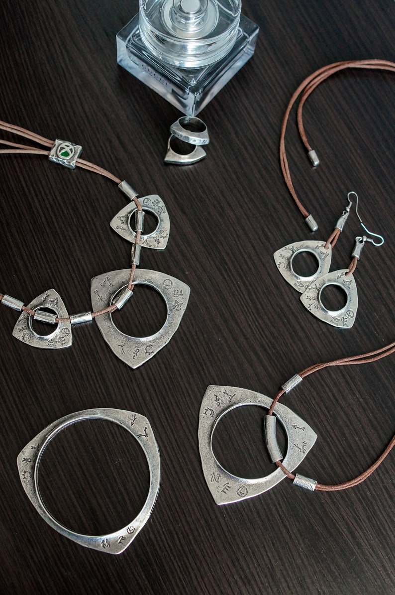 May include: A set of silver jewelry with a triangular pendant design. The pendants feature a circle with intricate engravings and are attached to brown leather cords. The set includes a necklace, earrings, and a bracelet.