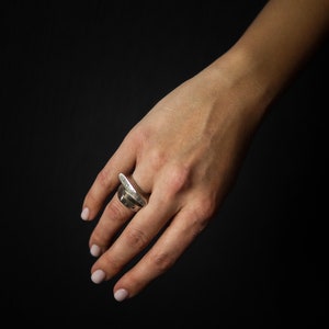 May include: A silver ring with a unique, organic design. The ring is worn on the middle finger of a hand with light pink nail polish.