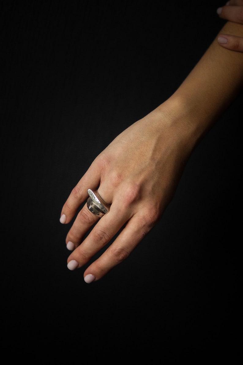 May include: A silver ring with a unique, organic design. The ring is worn on the middle finger of a hand with light pink nail polish.
