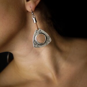 May include: Silver triangle earrings with a circular cutout and a brown leather cord. The earrings are hanging from a person's ear.