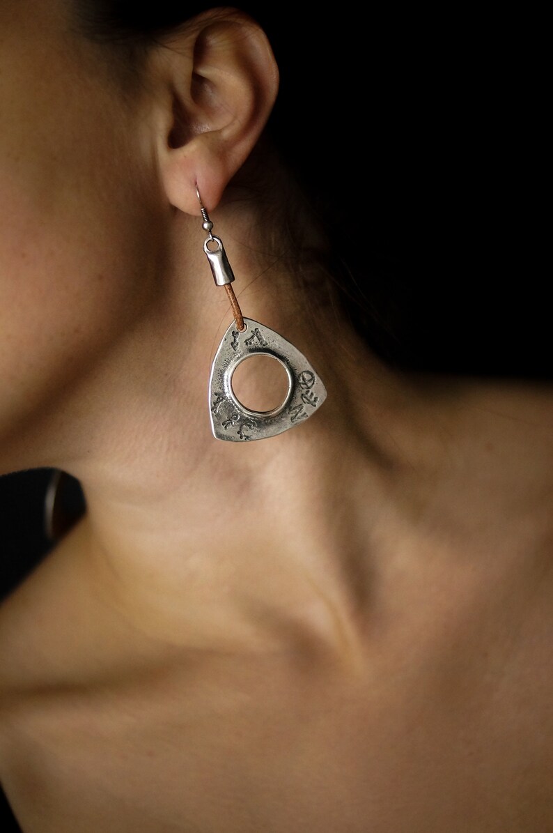 May include: Silver triangle earrings with a circular cutout and a brown leather cord. The earrings are hanging from a person's ear.