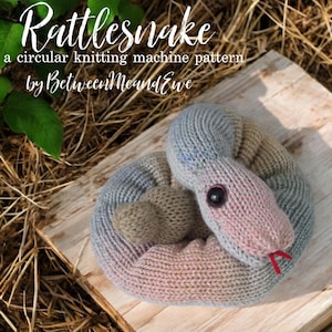 Op de afbeelding: Een gebreid rammelaar speelgoed in grijstinten, roze en bruin, opgerold op een houten oppervlak. De slang heeft een roze kop, een zwart knoopoog en een rode tong. De tekst "Rattlesnake" en andere tekst zijn zichtbaar.