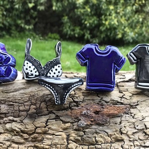 May include: Five ceramic figurines of different clothing items. A blue bra and panty set, a black bra and panty set, a blue t-shirt, and two t-shirts in black and white.
