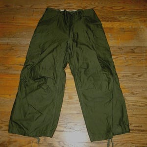 May include: Olive green military-style pants with a drawstring waist and pockets. The pants are made of a lightweight fabric and have drawstrings at the ankles. The pants are laid out on a wooden floor.