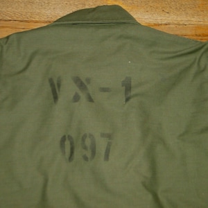 Peut inclure: Veste militaire vert olive avec le texte "VX-1 097" pochoir sur le dos. La veste a un col et un devant boutonné. Le tissu semble être un mélange durable, peut-être de coton.