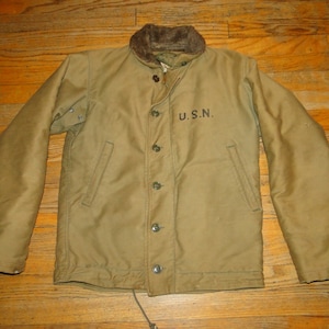 Peut inclure: Une veste kaki de style militaire avec un col doublé de fourrure et une taille à cordon. La veste a un patch U.S.N. sur la poitrine gauche.