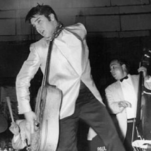 Peut inclure: Une photo en noir et blanc d'un homme jouant de la guitare sur scène. Il porte une veste de costume blanche et un pantalon noir. Un homme en smoking blanc joue de la basse derrière lui.