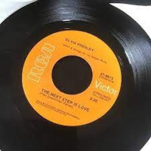 Puede incluir: Un disco de vinilo negro de 45 RPM con una etiqueta central naranja que presenta el texto Elvis Presley, The Next Step Is Love, RCA Victor, 47-9872 y 3:30. La etiqueta incluye el logotipo de RCA e indica que el disco fue hecho en los EE. UU.