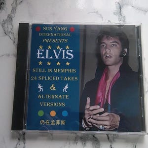 Peut inclure: Un boîtier de CD avec Elvis Presley. La couverture présente un design bleu et blanc avec le texte "SUN YANG INTERNATIONAL PRESENTS ELVIS STILL IN MEMPHIS 24 SPLICED TAKES & ALTERNATE VERSIONS". L'image de droite montre Elvis en manteau noir et écharpe rose.