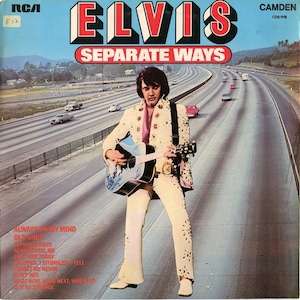 Puede incluir: Portada del álbum "Separate Ways" de Elvis Presley. La imagen muestra a Elvis con un traje color crema con recortes florales, sosteniendo una guitarra. El fondo muestra una autopista con coches. El título del álbum y el nombre del artista están en negrita, en rojo y blanco.