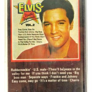 May include: A vintage Elvis Presley "Double Dynamite Vol. 2" cassette tape. The cover features a portrait of Elvis with the text "Elvis Double Dynamite" and a list of song titles. The cassette is in a clear plastic case.