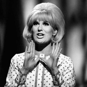 May include: Black and white photograph of a woman with blonde hair styled in a bouffant. She wears a polka dot blouse and has her hands raised. A ring is visible on her finger. The background is blurred.