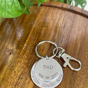 Puede incluir: Llavero de plata con dos etiquetas redondas de metal. La etiqueta superior dice "DAD" y la etiqueta inferior dice "we &hearts; you Sophie. Kevin. Amy".