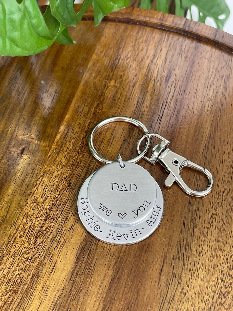 Puede incluir: Llavero de plata con dos etiquetas redondas. La etiqueta superior dice "DAD" y la etiqueta inferior dice "Sophie. Kevin. We &hearts; you. Amy".