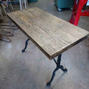 Peut inclure: Un plateau de table en bois rustique avec un piètement en métal noir. Le plateau de table a un motif de grain de bois brun foncé. Le piètement a un design décoratif avec trois pieds.