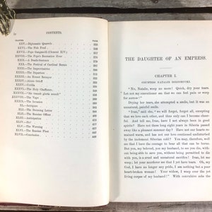 Puede incluir: Libro antiguo abierto con el t&iacute;tulo "The Daughter of an Empress" y texto visible. El libro tiene una cubierta roja y bordes dorados. Una mano decorativa de metal sujeta las p&aacute;ginas abiertas.