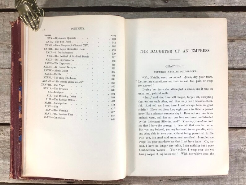 Puede incluir: Libro antiguo abierto con el t&iacute;tulo "The Daughter of an Empress" y texto visible. El libro tiene una cubierta roja y bordes dorados. Una mano decorativa de metal sujeta las p&aacute;ginas abiertas.