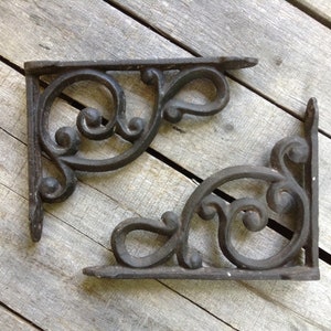 May include: Two ornate, dark brown cast iron shelf brackets with a scrolled design. The brackets are L-shaped, with a flat base and a decorative top. The brackets are placed on a weathered wooden surface.