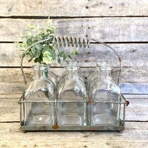 May include: A vintage-style glass bottle carrier with six clear square bottles. The bottles are held in a metal frame with a handle and a decorative spring. Faux greenery adds a touch of nature. The carrier is displayed on a weathered wooden surface.