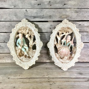 May include: Two vintage wall plaques with oval frames. One depicts a man in a blue coat, the other a woman on a swing. Both are cream-colored with detailed relief sculptures, perfect for adding a touch of classic elegance to any room.