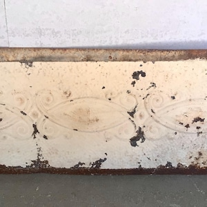 May include: Close-up of a section of a cream-colored, rectangular metal object with embossed scrollwork. The object has a distressed, aged appearance with areas of rust and paint chipping, revealing the metal underneath.