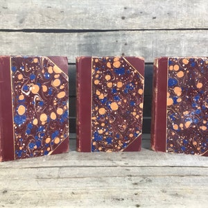 Puede incluir: Tres libros antiguos con cubiertas burdeos y dise&ntilde;os de papel veteado en tonos azules, naranjas y blancos. Los libros est&aacute;n de pie, mostrando sus lomos y cubiertas decorativas.