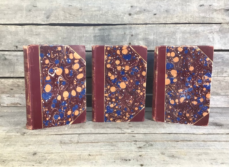 Puede incluir: Tres libros antiguos con cubiertas burdeos y dise&ntilde;os de papel veteado en tonos azules, naranjas y blancos. Los libros est&aacute;n de pie, mostrando sus lomos y cubiertas decorativas.