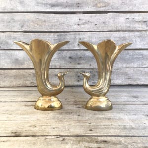 May include: Two gold-colored vases with a trumpet-like shape and a swan-like design. The vases have a wide, flared top and a curved body that tapers to a pedestal base. The swan's neck forms the handle of the vase.