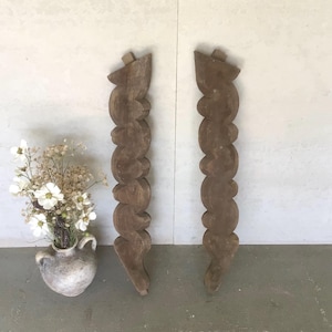 May include: Two antique, carved wooden architectural elements. The brown, vertical pieces have a spiral design and are likely decorative columns or posts. A small vase with white flowers is to the left.