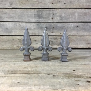 May include: Three gray metal fleur-de-lis fence finials. Each finial has a pointed top, a decorative base, and a square bottom. The finials are arranged in a row against a weathered wooden background.