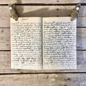 May include: An open vintage diary with handwritten cursive ink entries on two pages, held in place by two antique brass clips shaped like human hands with white pearl accents at the wrists, displaying detailed daily logs starting with April.