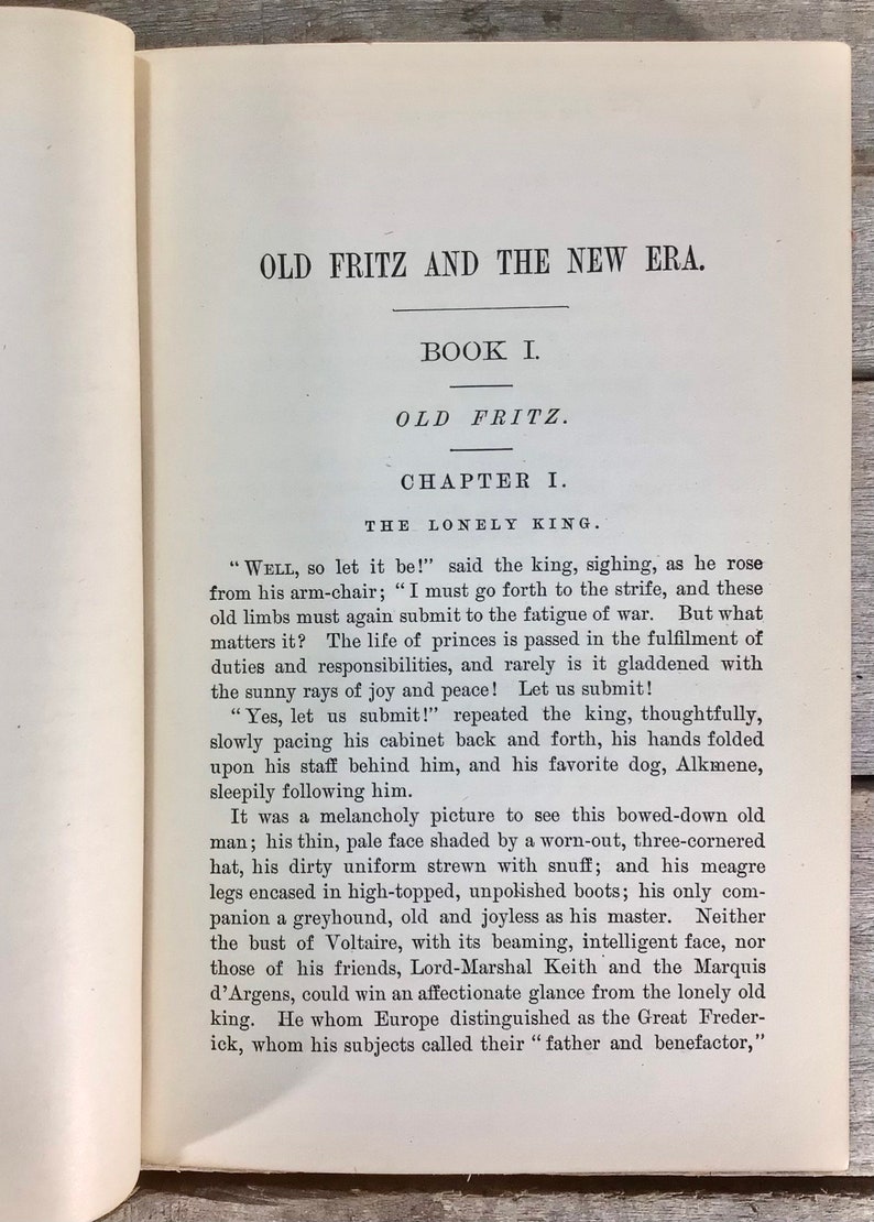 Puede incluir: P&aacute;gina de libro abierto con texto negro sobre papel blanquecino. El t&iacute;tulo es "OLD FRITZ AND THE NEW ERA." El texto contin&uacute;a con "BOOK I. OLD FRITZ. CHAPTER I. THE LONELY KING." El fondo es de madera.