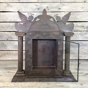 May include: A dark metal shrine cabinet featuring two flanking columns, a central rectangular opening with a hinged glass door, and a decorative pediment displaying two winged angels and an eye within a triangle symbol, all with a rustic finish.