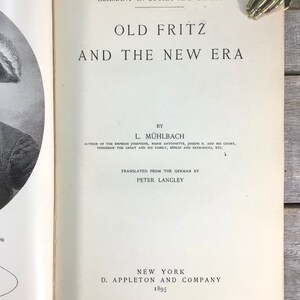 Puede incluir: Libro abierto titulado "Old Fritz and the New Era" de L. M&uuml;hlbach, publicado en 1895 por D. Appleton and Company, Nueva York. El t&iacute;tulo del libro est&aacute; en letras grandes y en negrita. El libro est&aacute; abierto en la portada.