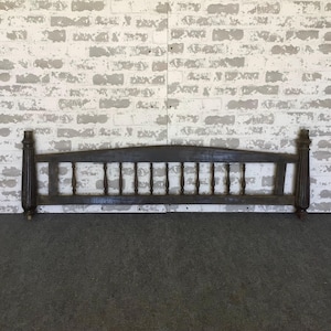 May include: Dark stained wooden headboard with a curved top and vertical spindles. The headboard has decorative posts on each end and is set against a white and gray brick wall. The headboard is a vintage piece.