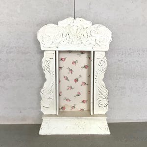 May include: A white, ornate, wooden wall shelf with a distressed finish. The shelf features a floral patterned backing and decorative scrollwork. The shelf is designed to hold small items and has a vintage aesthetic.