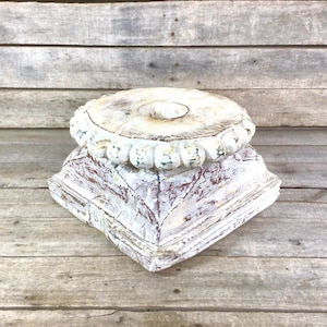 May include: A distressed, white-washed wooden pedestal with a square base and a round top. The top has a central hole and is surrounded by a decorative beaded edge. The wood shows red tones beneath the white paint.