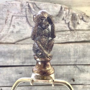 May include: Bronze-colored lamp finial featuring a seated monkey covering its eyes. The monkey is detailed with textured fur and is mounted on a decorative base. The finial is attached to a silver-colored lamp harp.