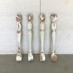 May include: Four antique wooden spindles with a distressed white paint finish. The spindles have a turned design with a square base and top. The wood shows through the paint in areas, giving them a rustic appearance. They are standing on a gray surface.
