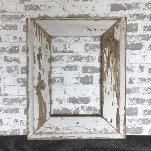 May include: A rectangular, distressed wooden picture frame with peeling white paint, revealing the natural wood underneath. The frame has a rustic, vintage aesthetic, perfect for displaying photos or artwork. The frame is empty.