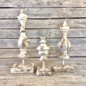 May include: Three distressed white decorative objects with gold accents. Each object has a unique shape, with a square base and a tall, ornate top. The objects are arranged against a weathered wood background.