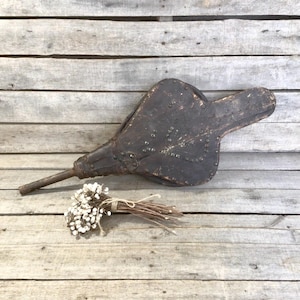 May include: A vintage, dark brown leather bellows with a metal nozzle and decorative rivets. A small bundle of dried white flowers tied with twine rests nearby. The bellows is likely used for a fireplace or forge.