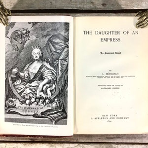 Puede incluir: Un libro abierto titulado "THE DAUGHTER OF AN EMPRESS" con una ilustraci&oacute;n de una mujer con vestimenta hist&oacute;rica. El libro est&aacute; abierto por dos sujetalibros decorativos en forma de mano. La portada del libro es de color burdeos.