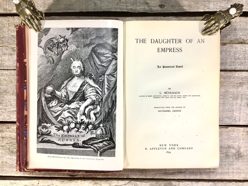 Puede incluir: Un libro abierto titulado "THE DAUGHTER OF AN EMPRESS" con una ilustraci&oacute;n de una mujer con vestimenta hist&oacute;rica. El libro est&aacute; abierto por dos sujetalibros decorativos en forma de mano. La portada del libro es de color burdeos.