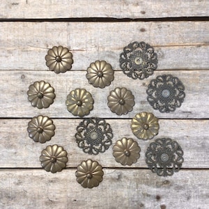 May include: An assortment of antique brass-toned drawer pulls and decorative metal accents. The drawer pulls have a flower-like design, while the accents feature ornate, openwork patterns. The items are arranged on a weathered wooden surface.