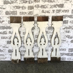 May include: Four vintage white wooden balusters with decorative cutouts and brown tops. The balusters are arranged against a distressed white brick wall, showcasing their aged appearance and unique design. The balusters are approximately 30 inches tall.